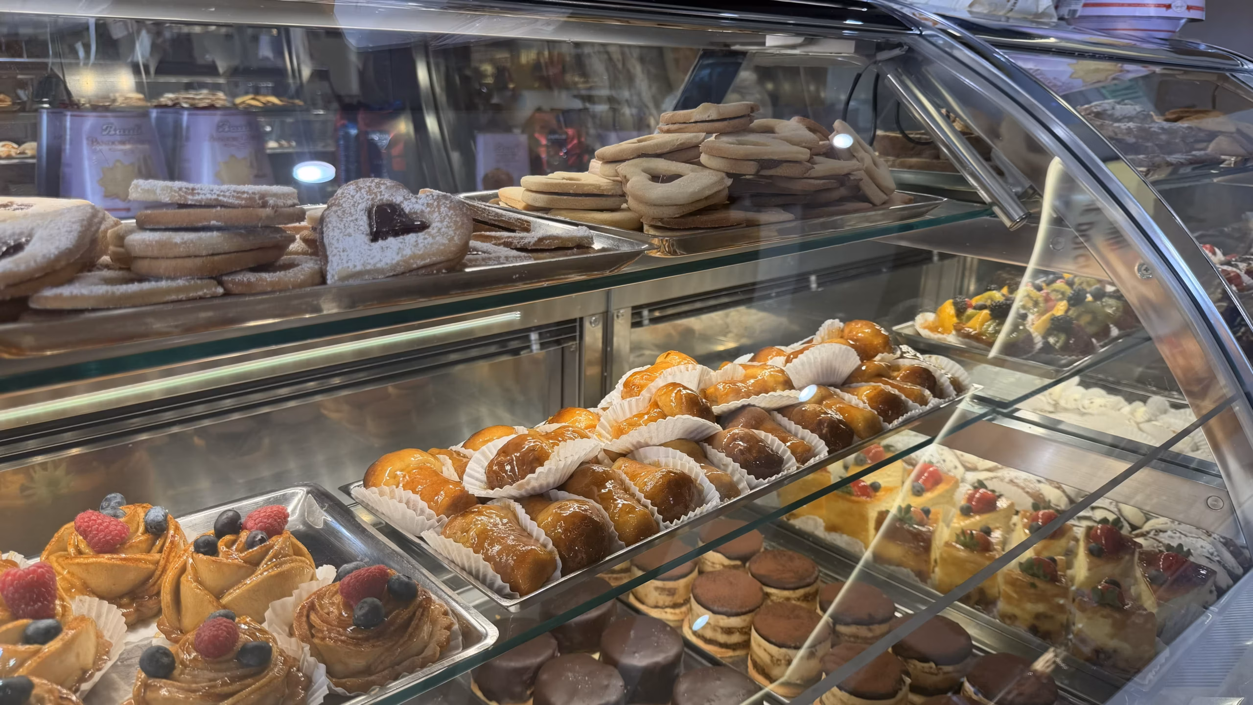 Glass display case filled with fresh Italian pastries, glazed brioche buns, fruit tarts, cookies, and cakes at an Italian bakery in Lauderdale-by-the-Sea