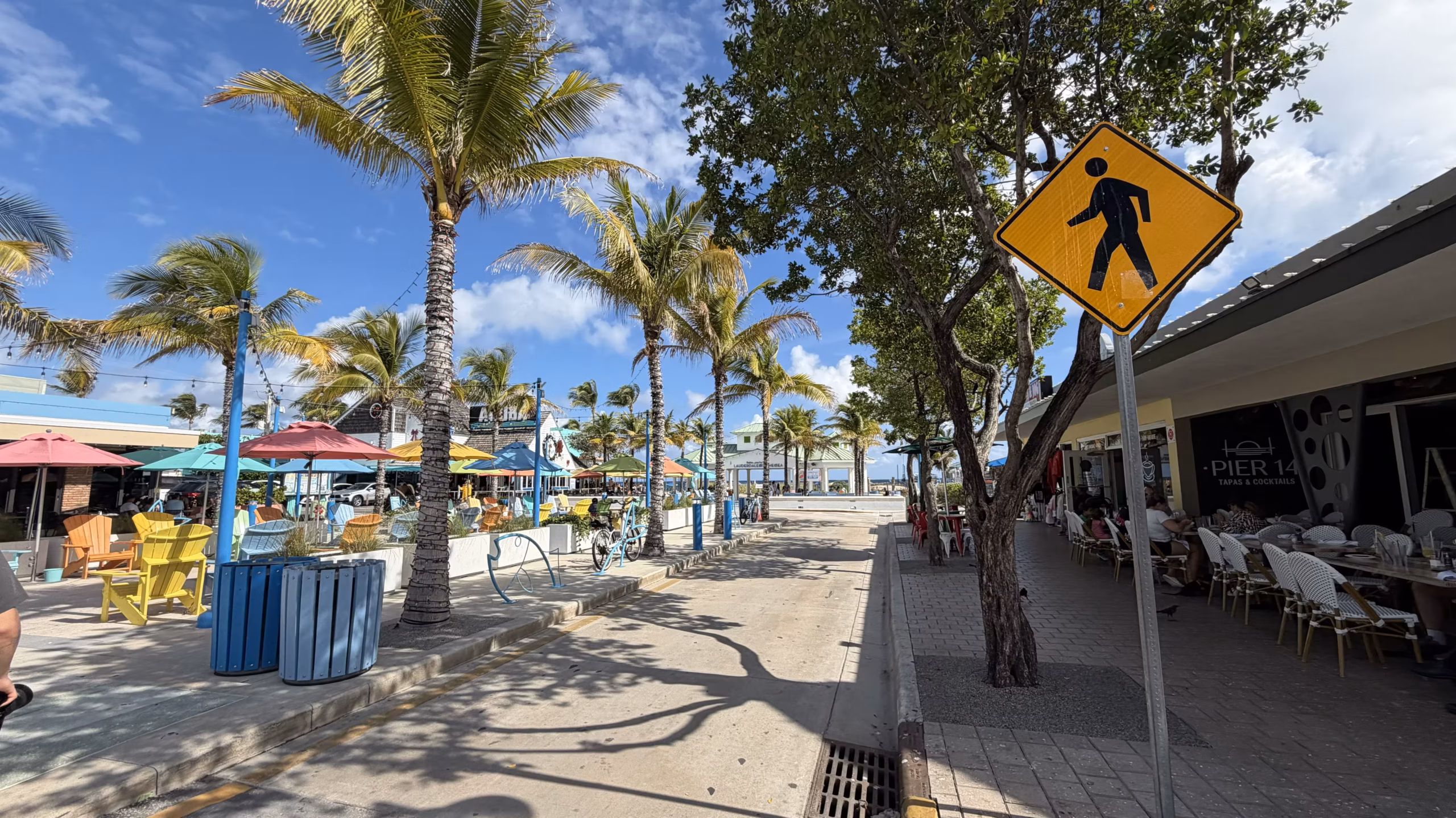 Pedestrian walkway in Lauderdale-by-the-Sea village with colorful restaurant terraces, palm trees, and outdoor seating along the main street